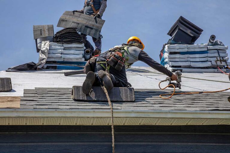 Homeowners inspecting a light-colored shingle roof under clear summer skies, surrounded by leafy residential streets.